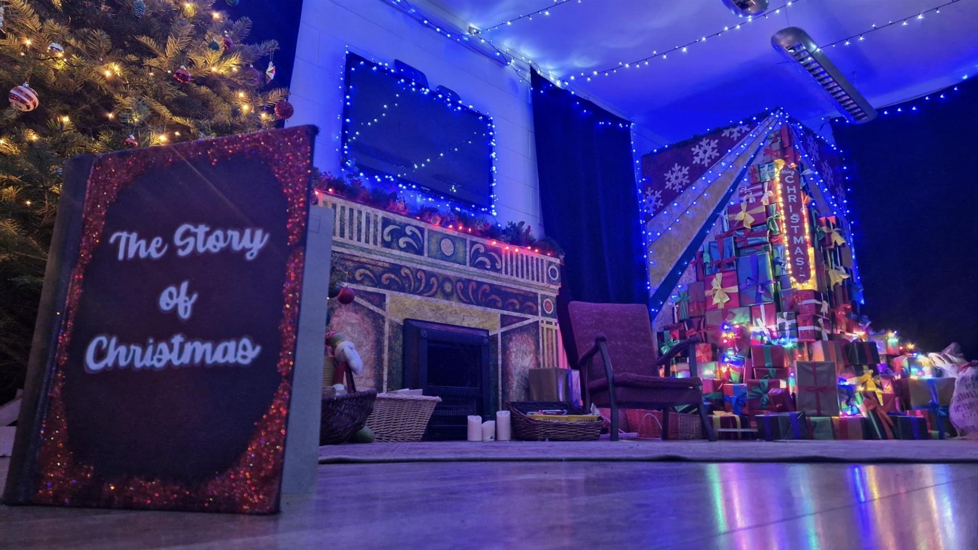 a quiet living room scene is depicted lit up with Christmas lights and a tree. a book is propped up on the floor and the front cover reads 'The Story