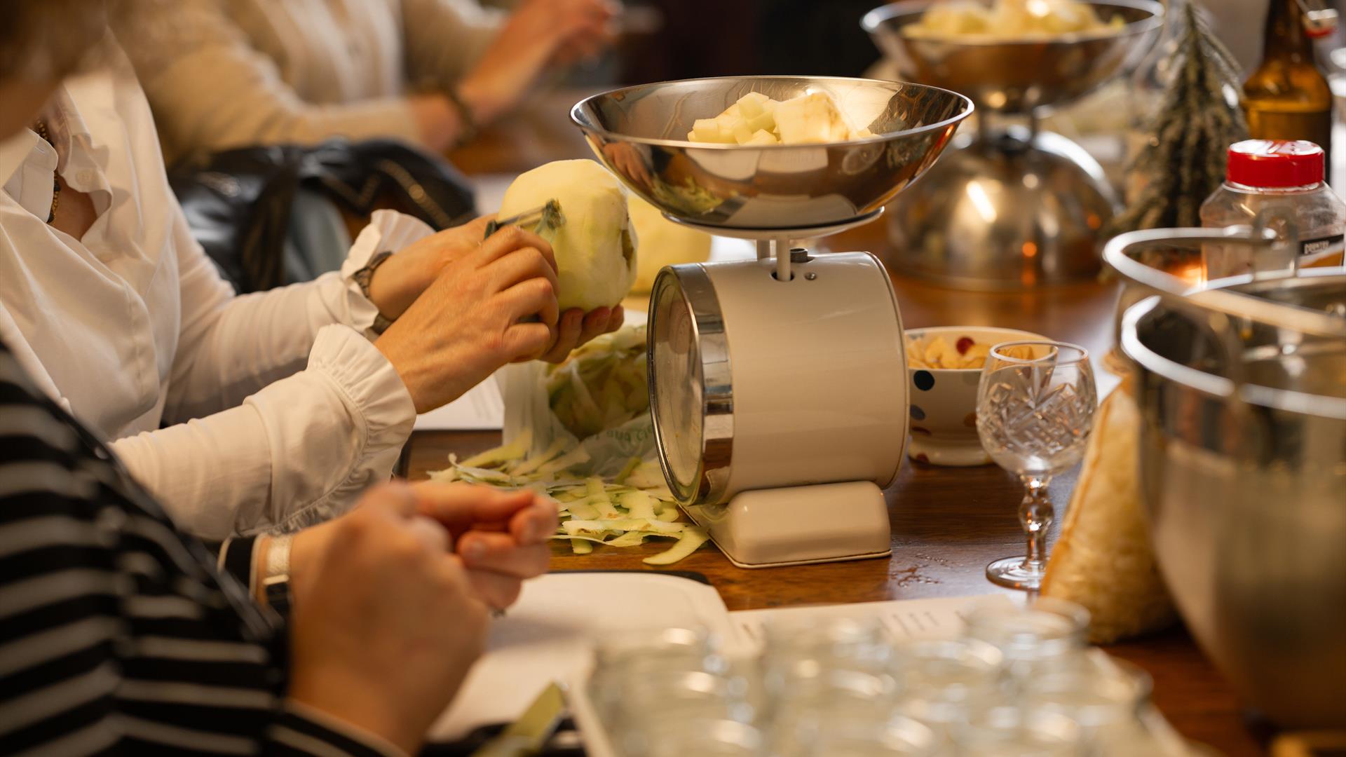 a person's hands peel a large cooking apple while a set of kitchen scales sits on the table infront of them