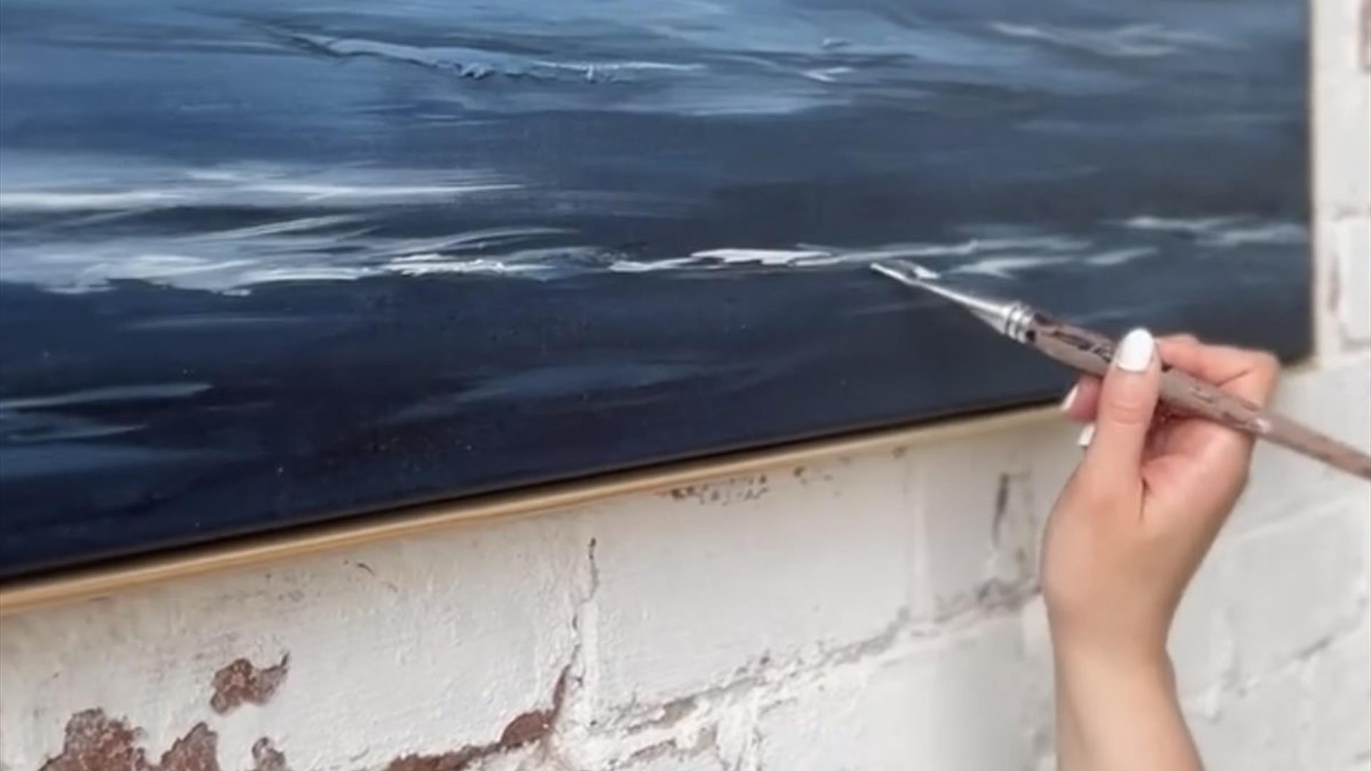 a hand holds a brush up to add finishing touches to a deep blue abstract painting of the sea