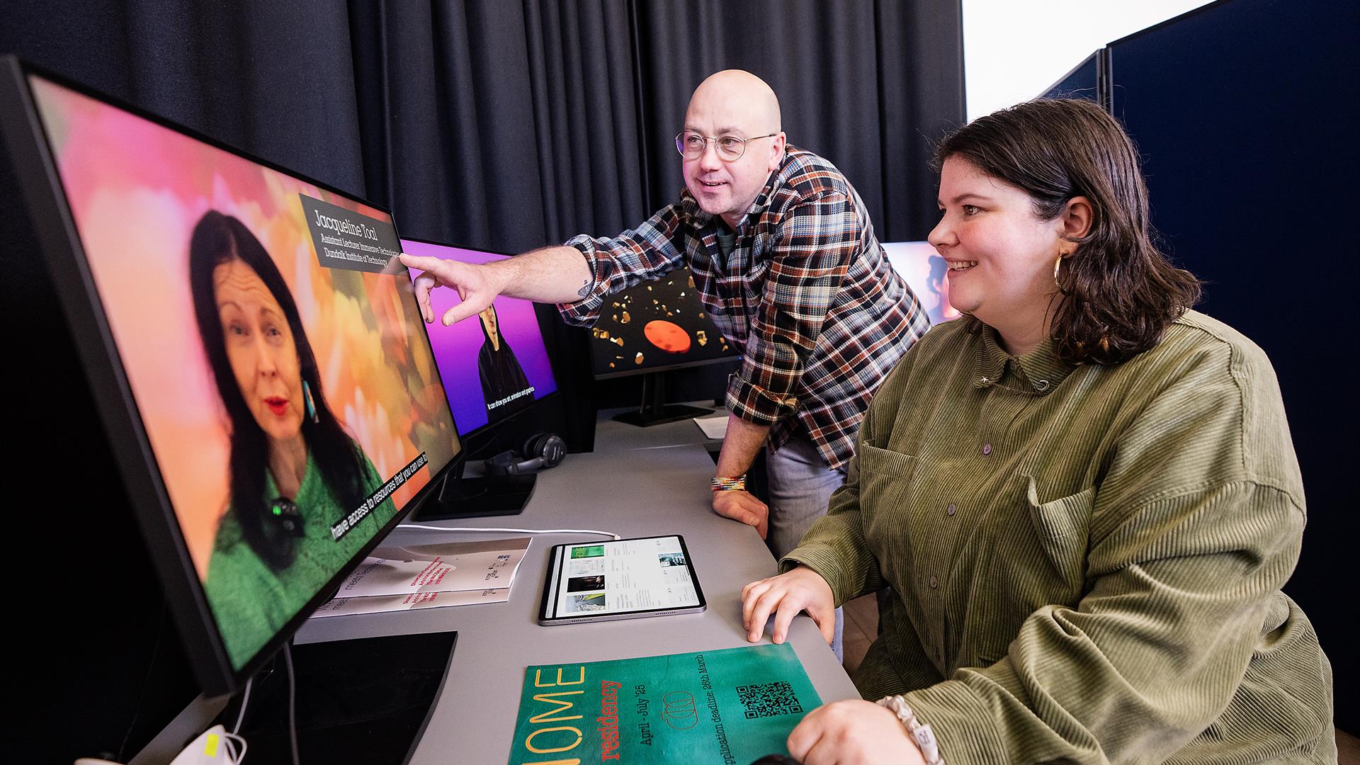 a man in glasses points towards a computer screen. next to him, a woman smiles