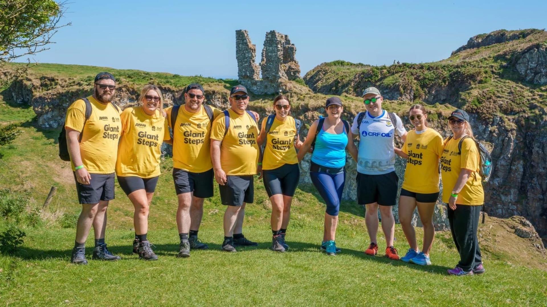 A group of participants in yellow Giant Steps t-shirts standing together on a grassy cliffside with coastal rock formations in the background, smiling