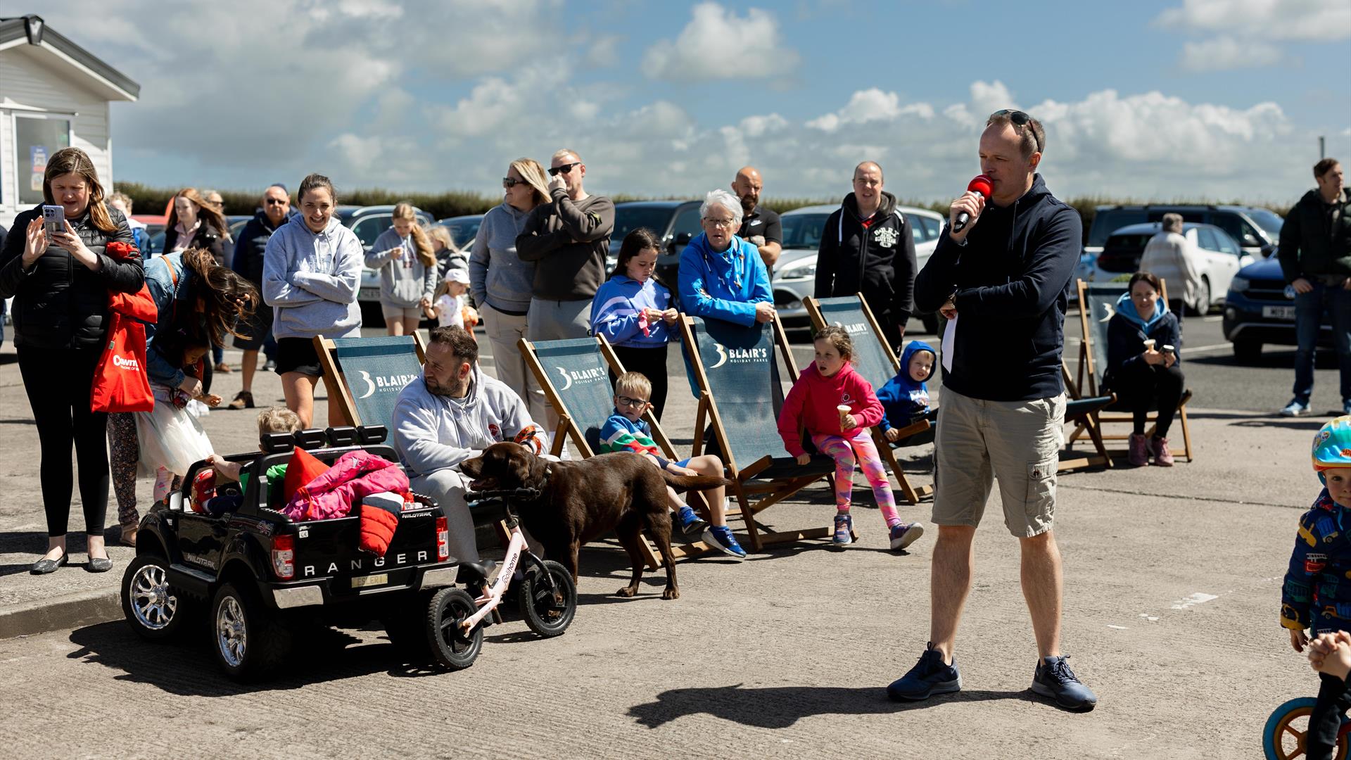 A family lounges on deck chairs surrounded by other families all taking pictures together and enjoying a day out, with a blue sky in the background.