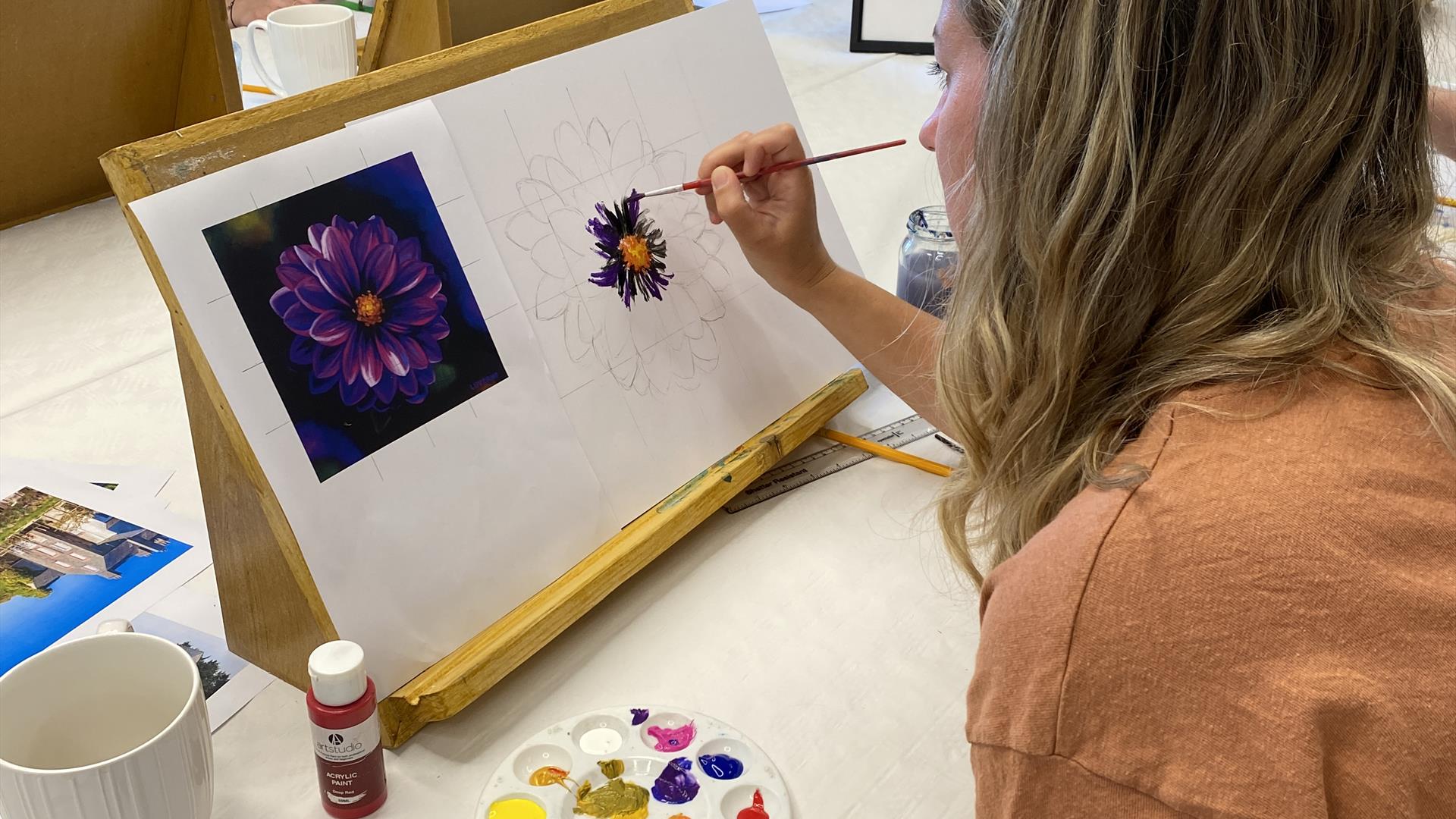 A woman painting a purple flower using a reference image attached to her canvas.