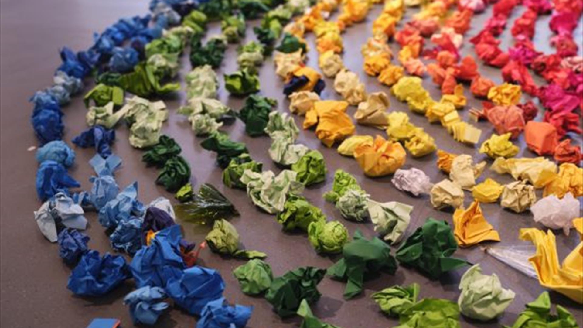 A number of crumpled pieces of coloured paper form a larger rainbow, in a circular shape.