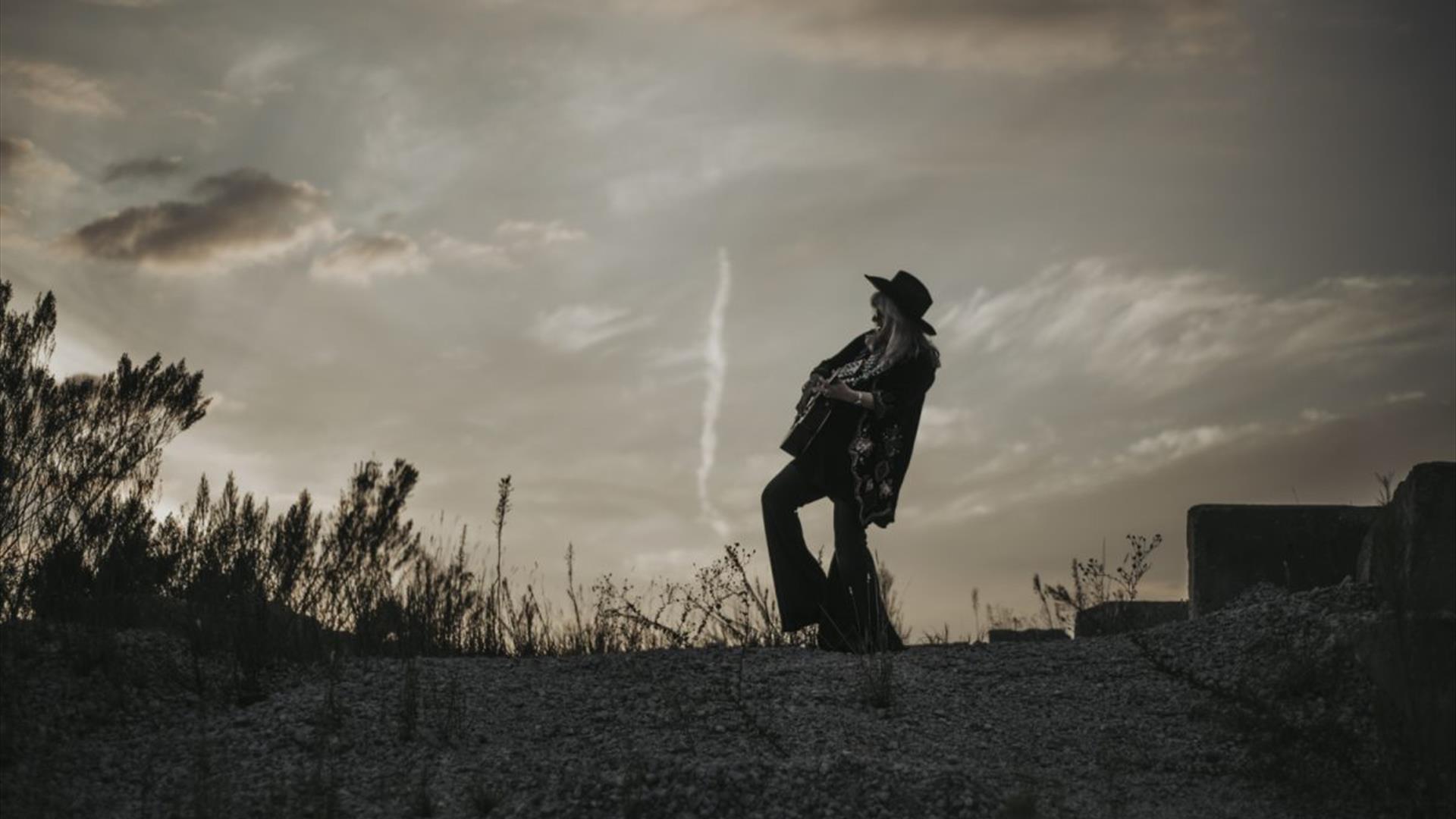 black and white image of kimmi rhodes playing a guitar silhoutted against the sky,