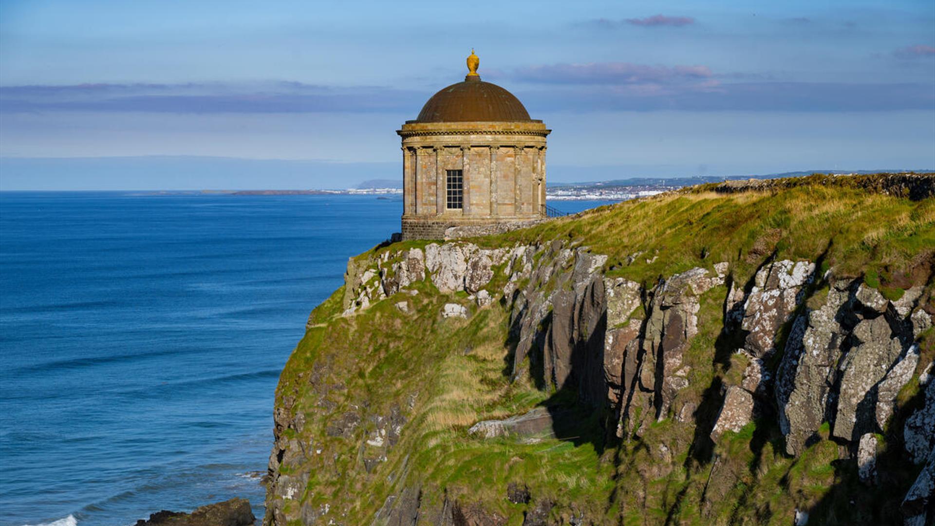 View of Mussenden Temple perched on a dramatic cliff edge above the Atlantic Ocean, seen along the coastal walking trail from Castlerock.