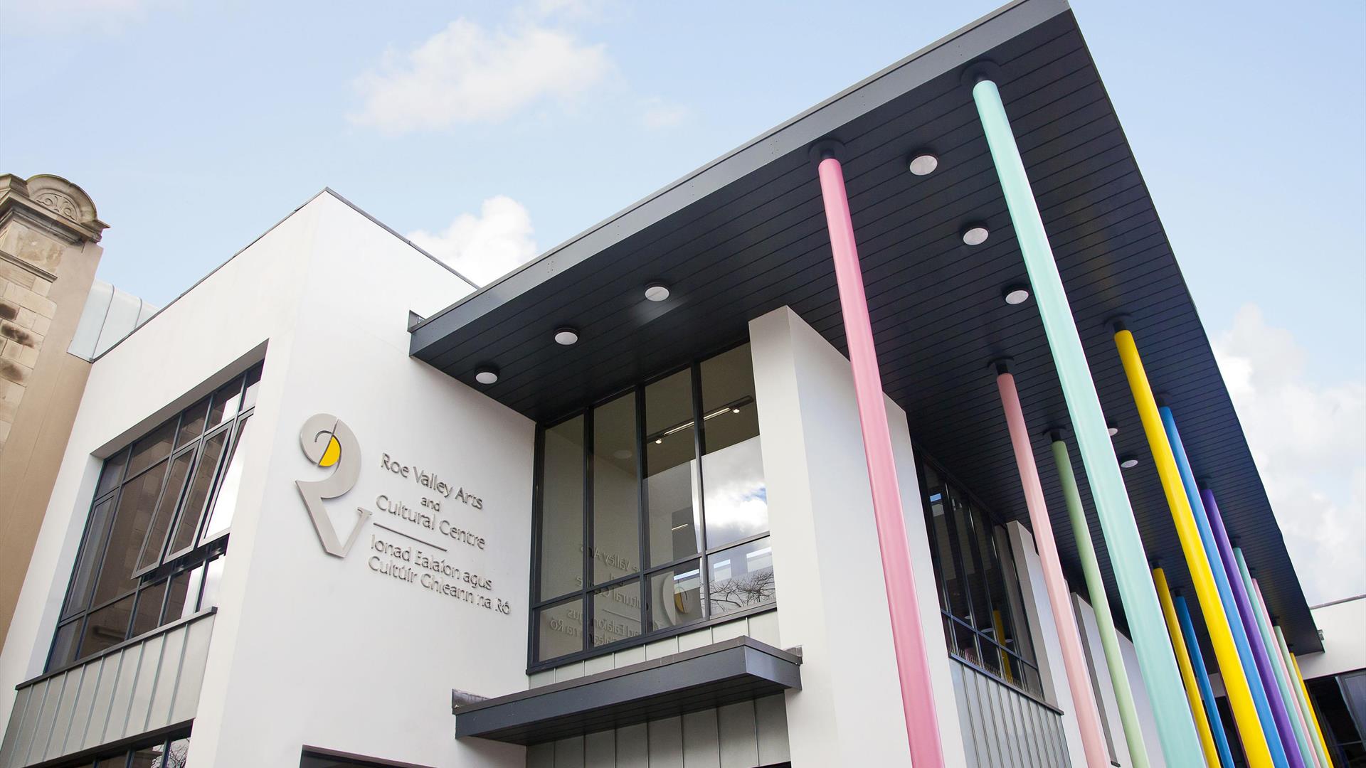 Outside view of the Roe Valley Arts & Cultural Centre - a white building with large windows and bright pastel coloured pillars at the side