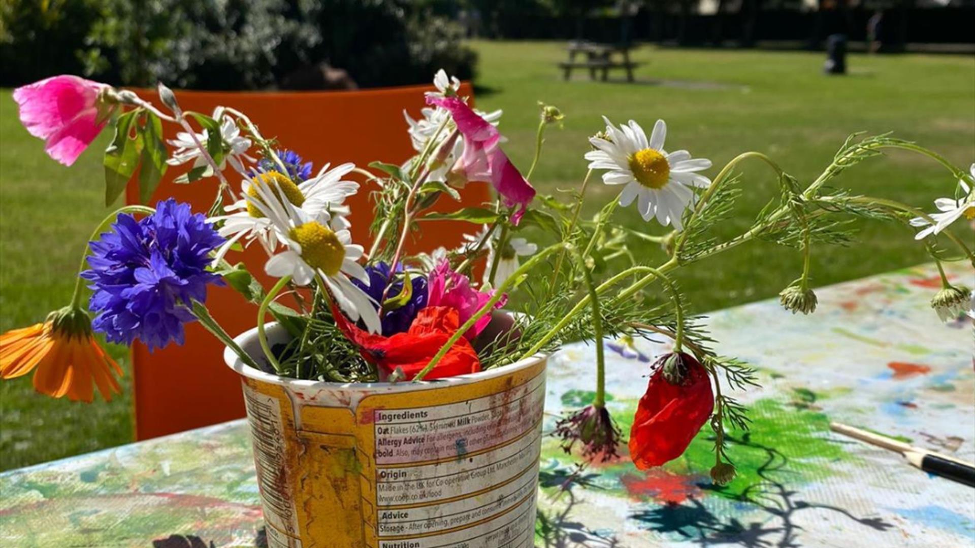 A bundle of colourful flowers potted in a makeshift planter, with a park and clear blue sky visible in the background.