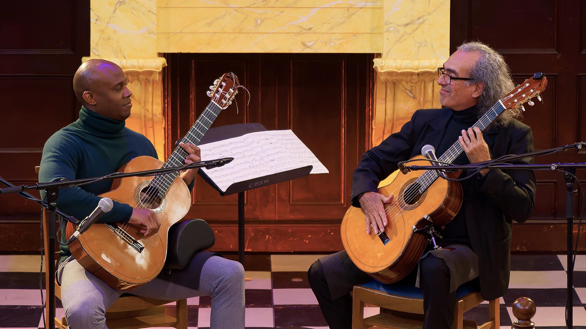 Two men hold their guitars while looking to each other with a smile. These are the members of The Bridge.