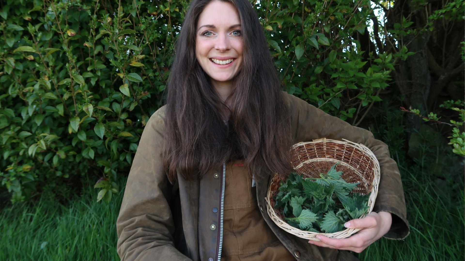 A woman stands smiling in the forest with a wicker basket full of foraged plants.