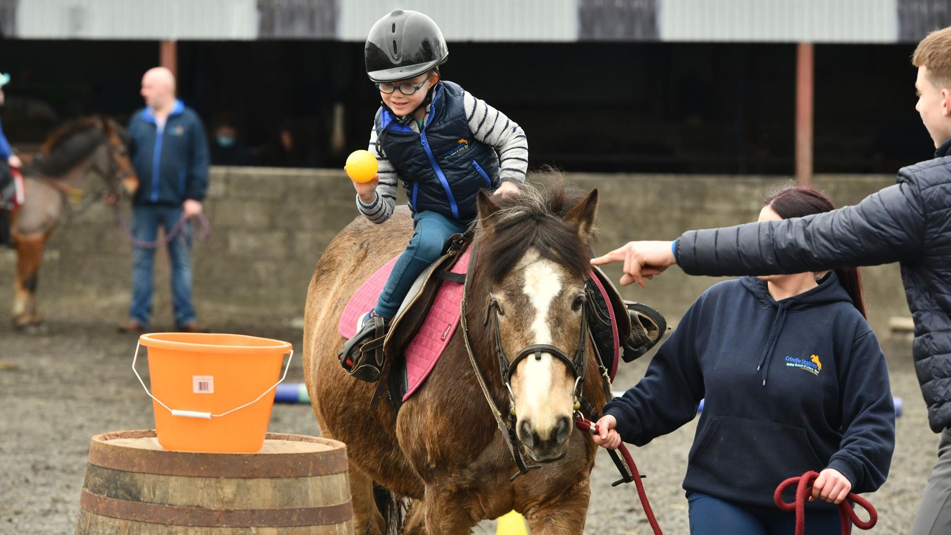 a child aims a ball towards a bucket while on horseback as Crindle Stables' instructors lead the horse