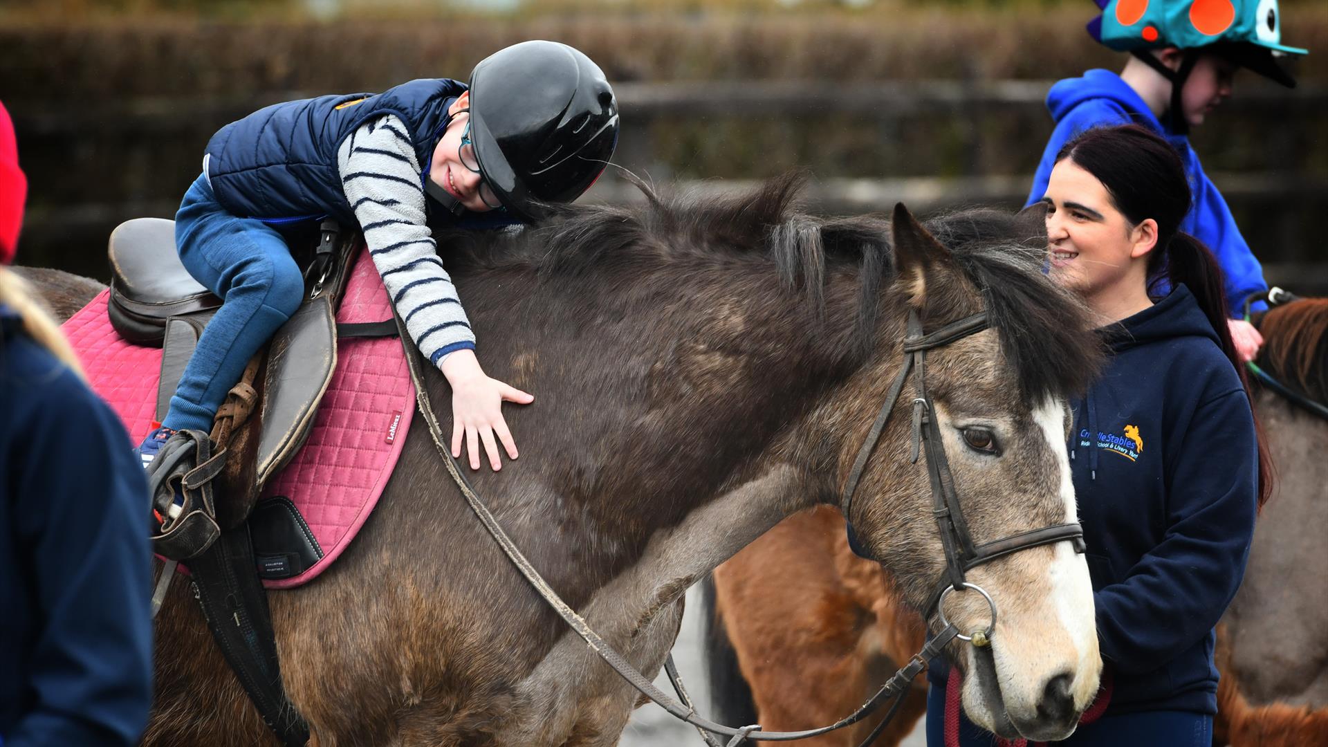 a child on horseback leans forward to hug a horse, while an instructor watches on