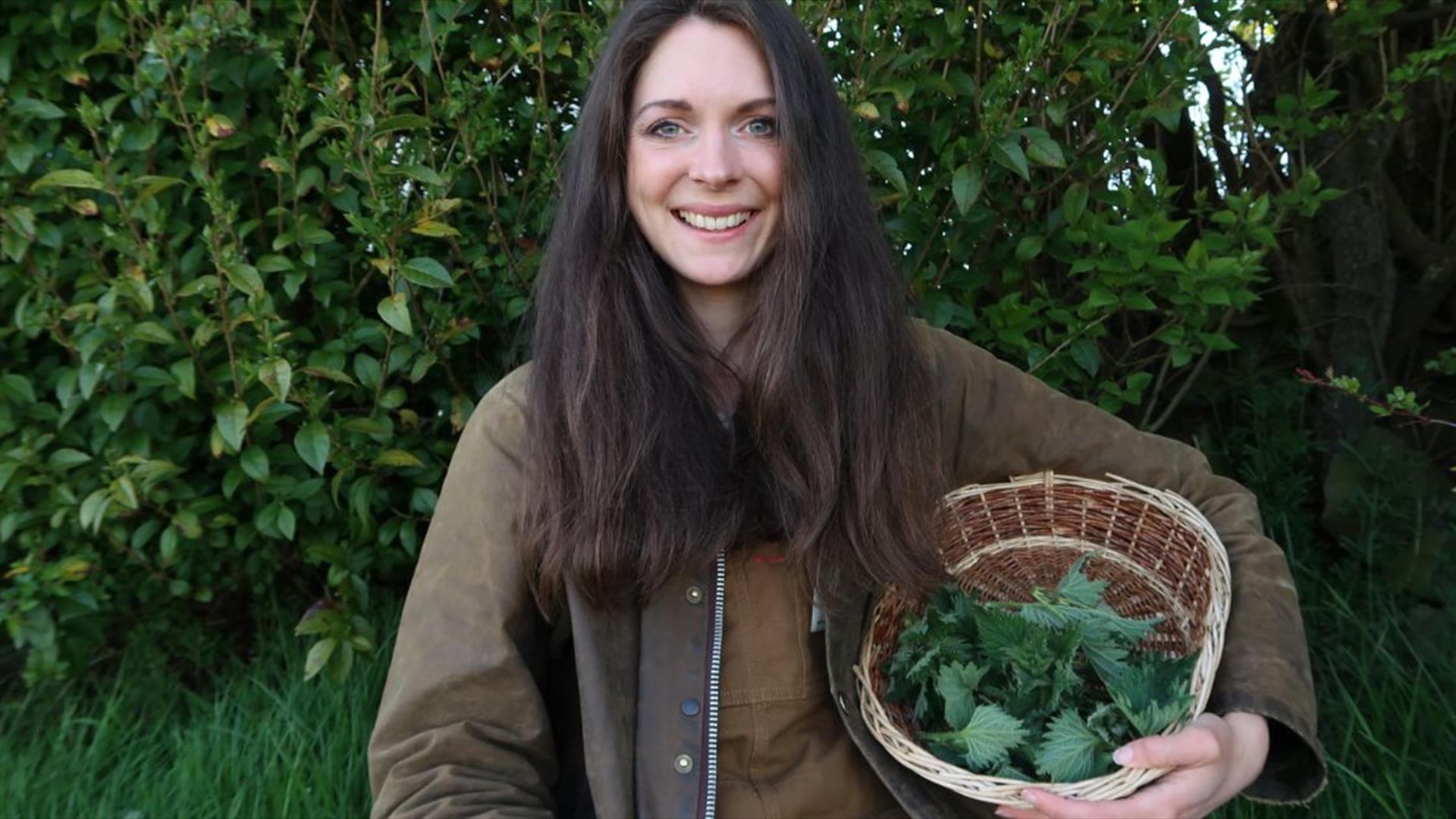 a woman holds a wicker basket containing foraged plants