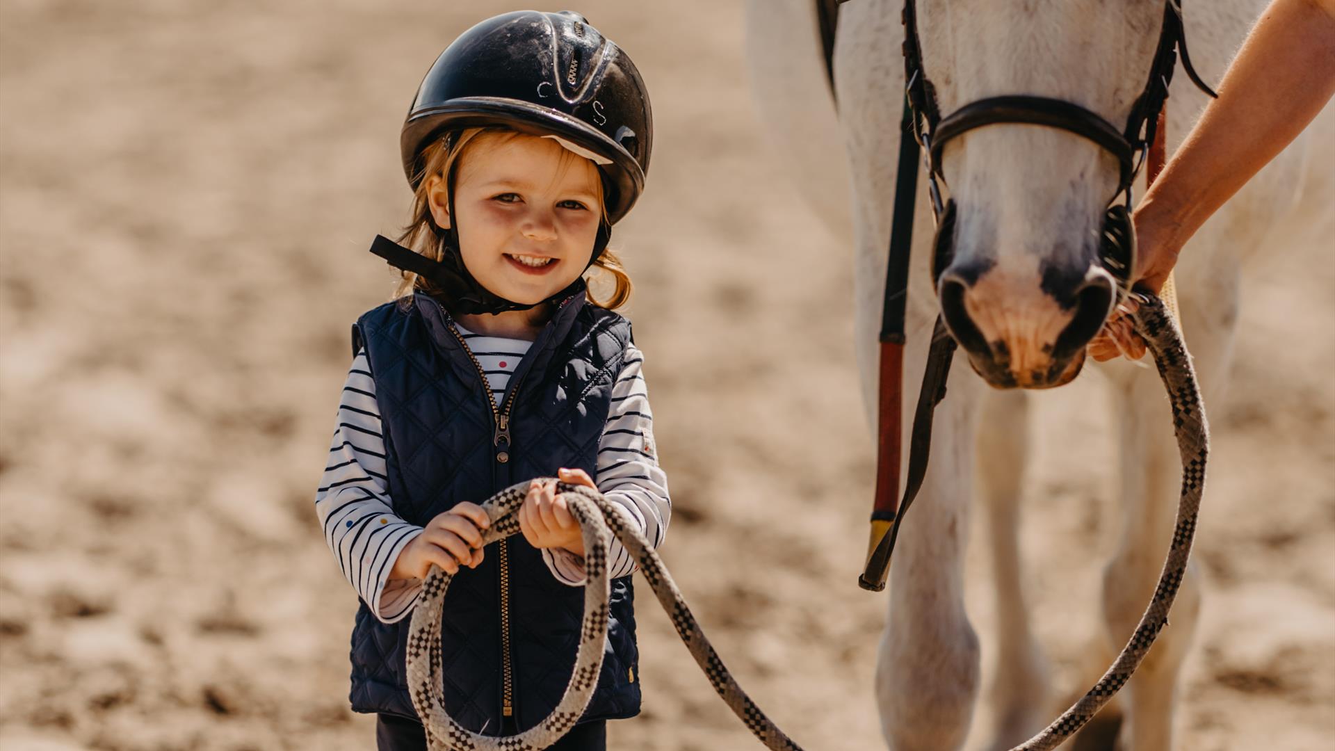 A little girl with a helmet on holding the reins of a white pony
