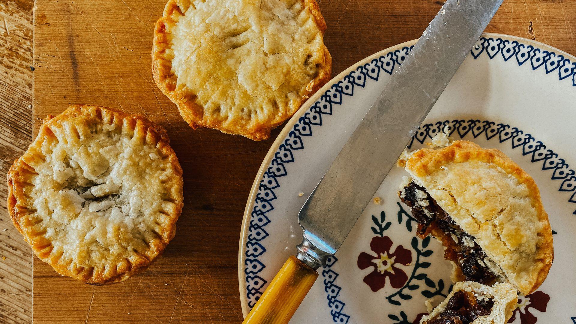 Image shows a mince pie cut open on a white and blue plate, with a knife beside it. There are also two mince pies on the table beside the plate.