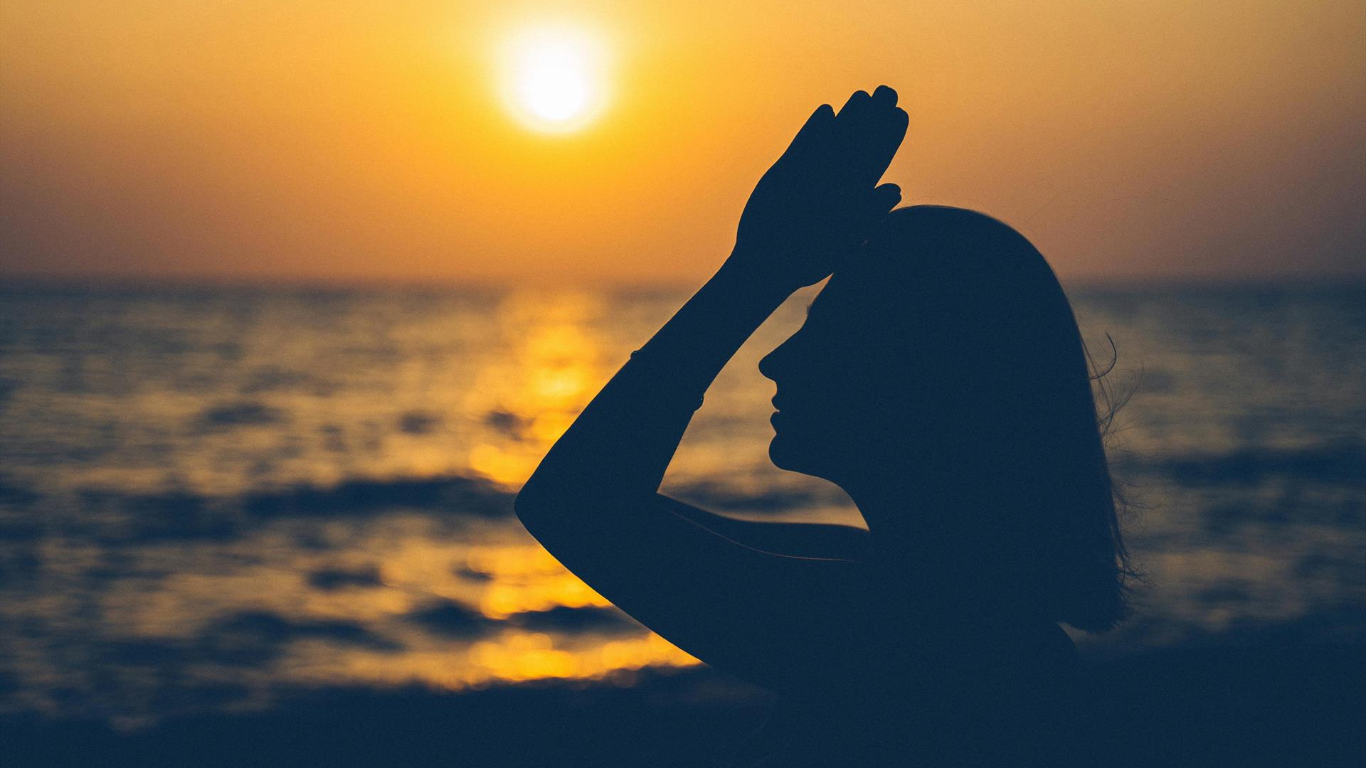 a person raises joined hands to their forehead with the sunset and the sea in the background