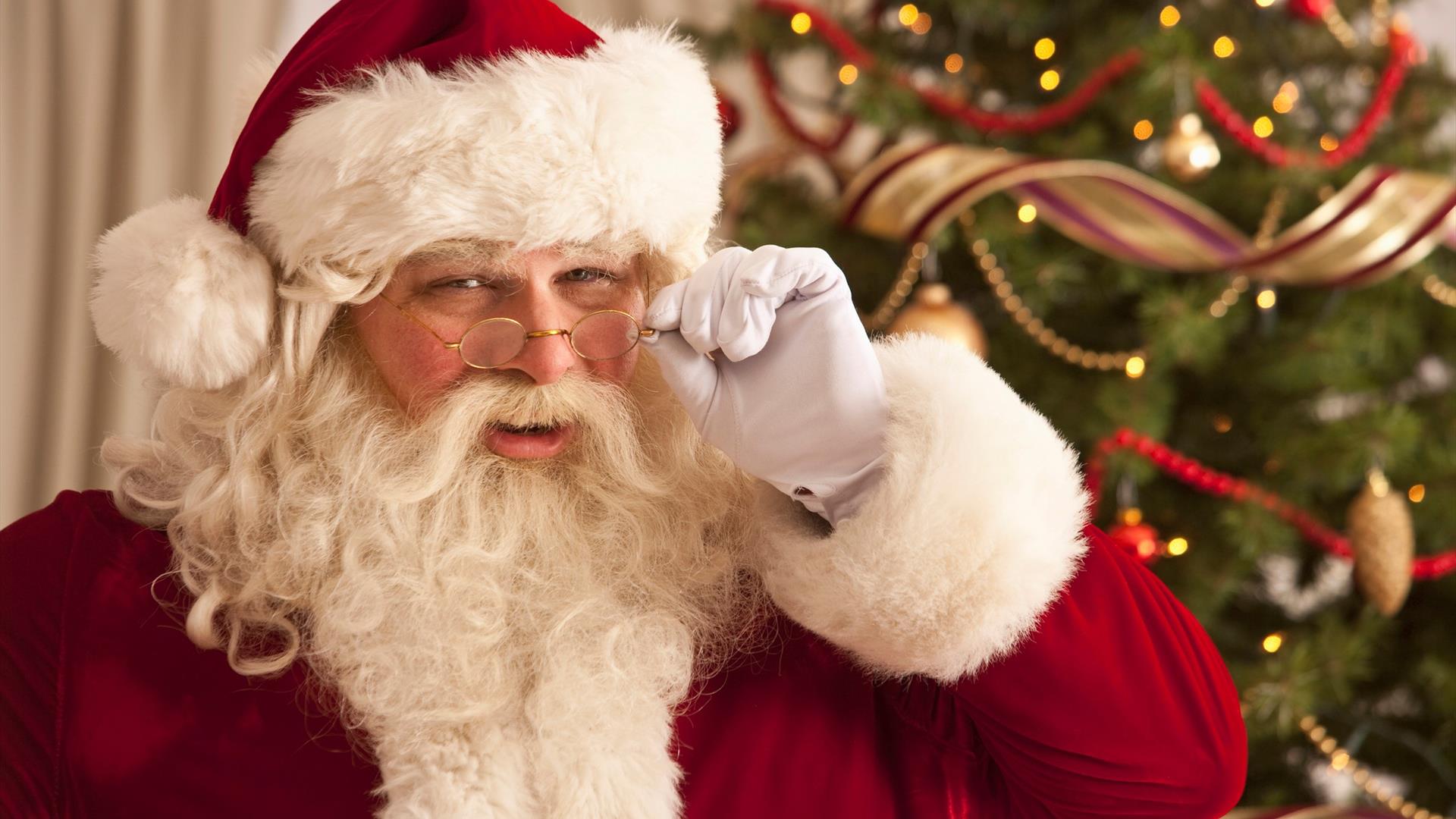 Santa peering over the rim of his glasses with a christmas tree in the background