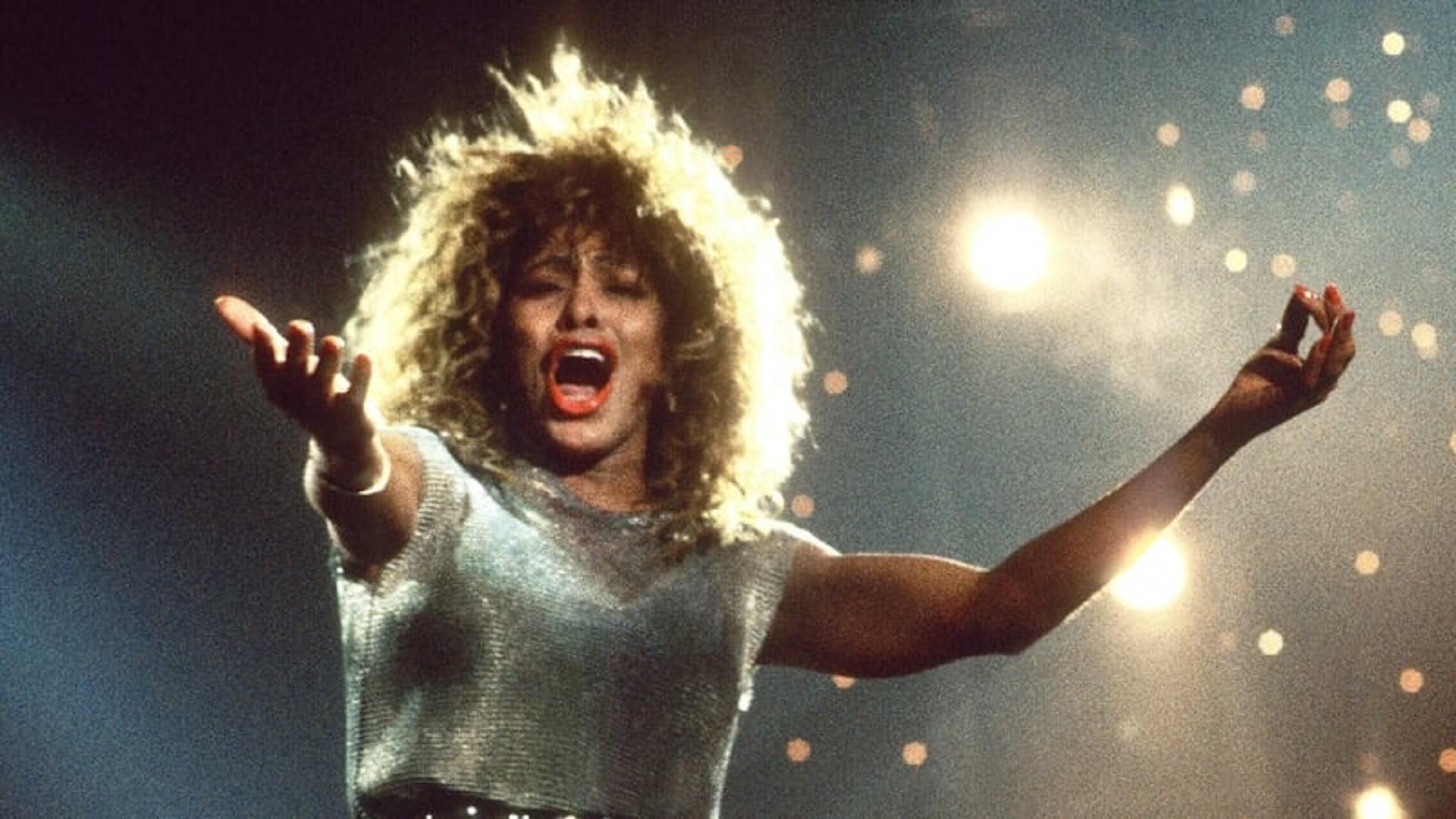 Tina Turner singing against sparkling lights with arms outstretched