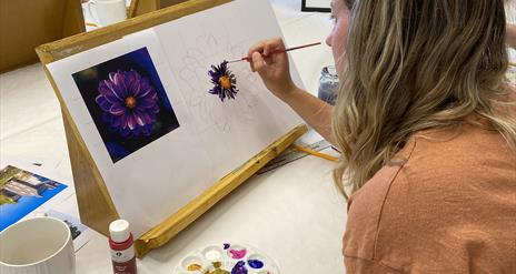 A woman painting a purple flower using a reference image attached to her canvas.