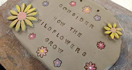 A ceramic sign with flowers carved into and embellishing it. It reads "Consider how the Wild Flowers grow"