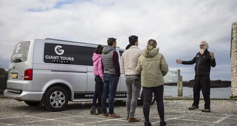 tour guide, Flip, talks to a group of tourists standing beside a van branded with the 'Giant Tours' logo