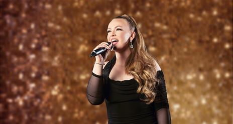 Image shows a woman wearing a black dress, singing into a microphone in her hand and standing in front of a gold glitter wall
