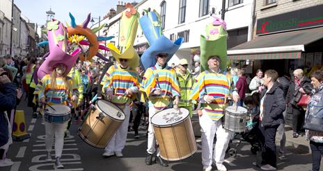 Carnival parade makes its way down the street with performers dressed in bright yellow costumes wearing tall coloured hats
