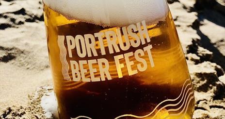 A glass of frothing beer on the sand. Text on the glass reads "Portrush Beer Fest"