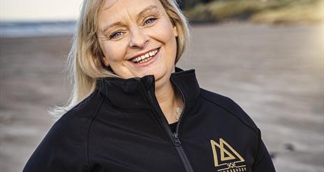 Claire O'Kane of Mussenden Sea Salt stands smiling beneath Mussenden Temple, wearing a black zip up fleece with her company logo in gold.