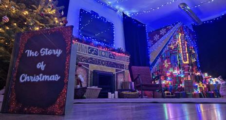 a quiet living room scene is depicted lit up with Christmas lights and a tree. a book is propped up on the floor and the front cover reads 'The Story