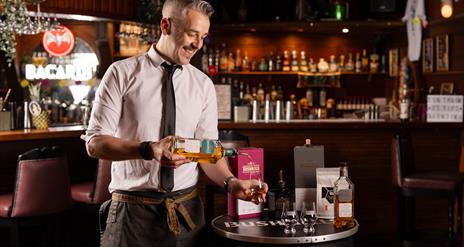 Bartender and mixologist, Ashely, pours whiskey in to a glass in front of a bar.