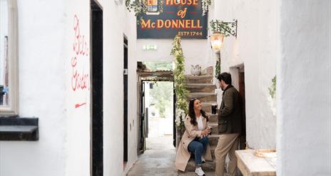 a woman sits on the steps at House of McDonnell while talking to a man who is standing, they are both holding drinks