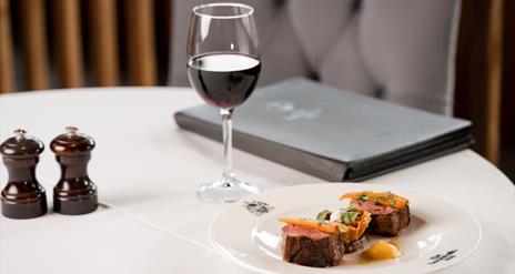 Image shows a chair and dining table covered in a white cloth, with a plate of food, a glass of red wine, a menu and salt/pepper grinders