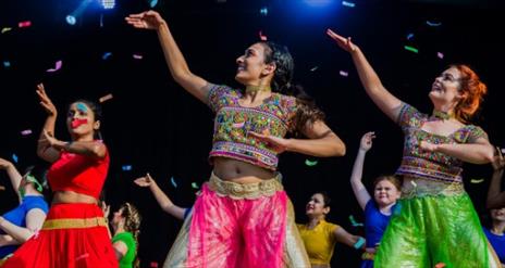 Image shows three female dancers wearing brightly coloured clothes and with one hand up in the air