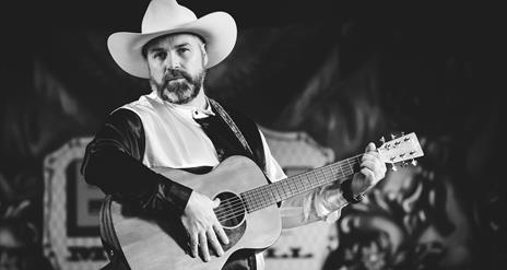 Black and white photograph o Stephen Butler with a guitar in a cowboy hat.