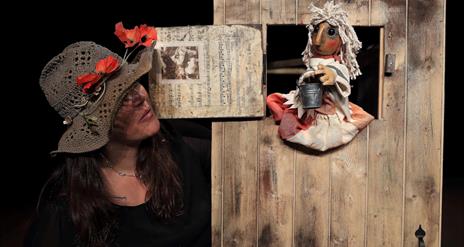 Person in a hat with red flowers on it standing next to a wooden door with a small hatch at the top through which a wooden puppet is sitting, hold a s