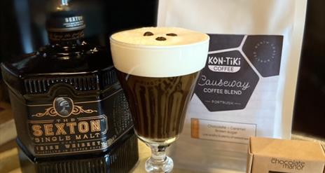 a bottle of Sexton Single Malt whiskey, an Irish Coffee with 3 coffee beans floating on the cream head, a bag of Kon-Tiki Causeway Coffee