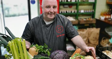 a man stands with a box filled with fresh vegetables including carrots, parsnips, celery, onions and cabbage