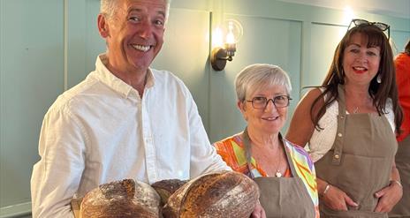 A man stands smiling holding loaves of fresh sourdough bread. Two women in aprons stand behind him and to the right.