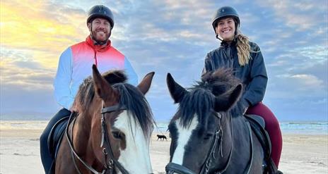 a man and woman sitting on horse back on the beach