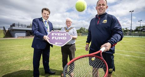 2 men hold a sign saying 'Event Fund' while another bounces a tennis ball on a racket