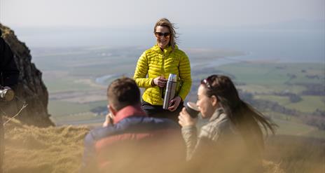 a man and woman sitting on a cliff drinking from cups. a woman stands in front of them with a thermal flask