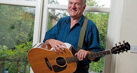 Image of a man in a blue shirt holding a guitar with a strap