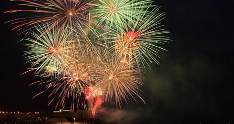 Fireworks burst to life above the sea and rocks, with the college in the background