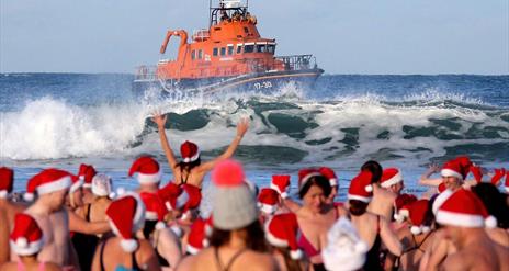 people in santa hats and swimwear watch on as a RNLI lifeboat cruises on the waves