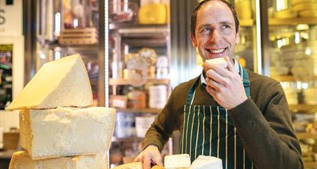 Kevin Sheridan in a striped apron, smiles while tasting cheese