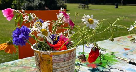 A bundle of colourful flowers potted in a makeshift planter, with a park and clear blue sky visible in the background.