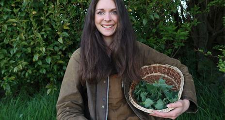 A woman stands smiling in the forest with a wicker basket full of foraged plants.