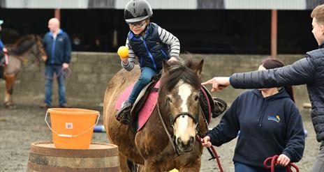 a child aims a ball towards a bucket while on horseback as Crindle Stables' instructors lead the horse