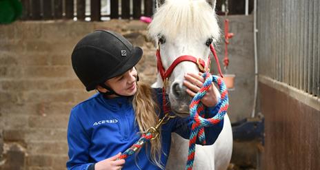Image shows a young girl in a blue top and black riding helmet petting a white horse
